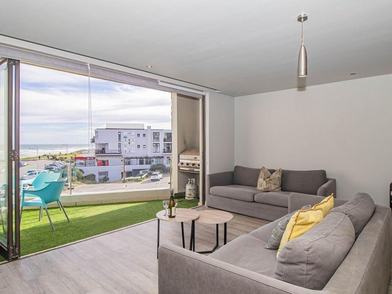 Open-plan living area featuring laminate flooring and large bi-fold doors opening onto a balcony with artificial grass and coastal views.