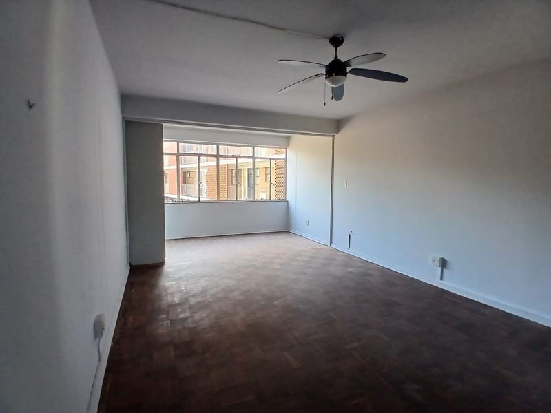 An empty living room featuring parquet flooring, a ceiling fan, and a large window providing natural light.