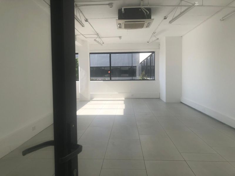 A bright, open-plan office space featuring tiled flooring, large windows for natural light, and a ceiling-mounted air conditioning unit.