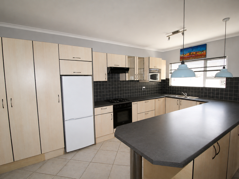 A functional kitchen featuring light wood-finish cabinetry, dark laminate countertops, a black tiled backsplash, and integrated appliances including an oven and hob.