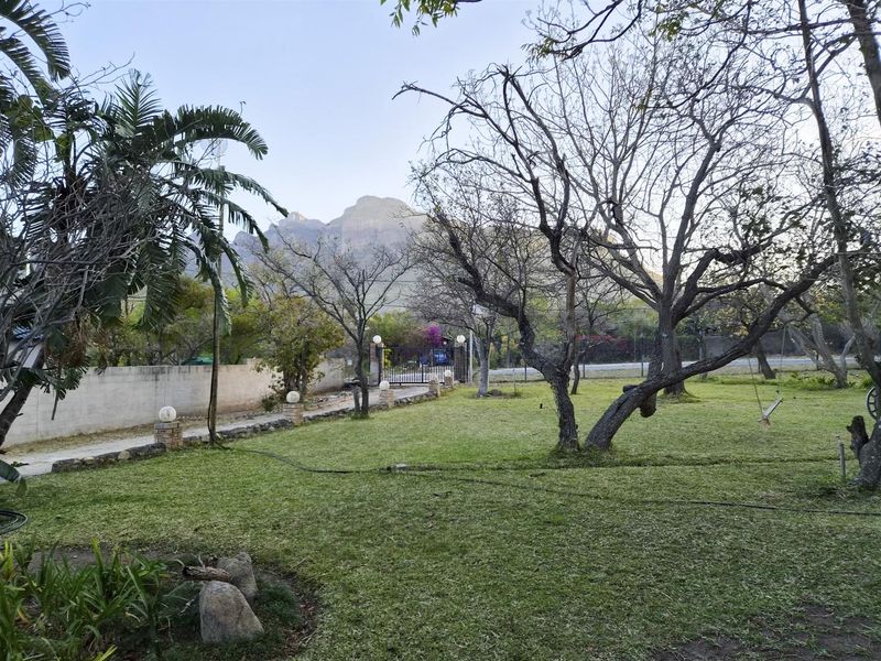 A spacious garden area featuring mature trees, a paved pathway, boundary wall, and a metal entrance gate with mountain views in the background.