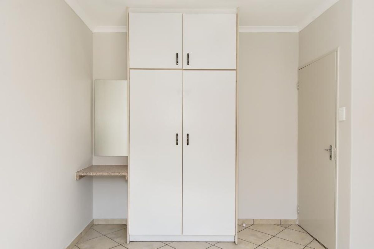 A clean, neutral bedroom featuring a built-in wardrobe with overhead storage and an integrated vanity shelf, finished with tiled flooring.