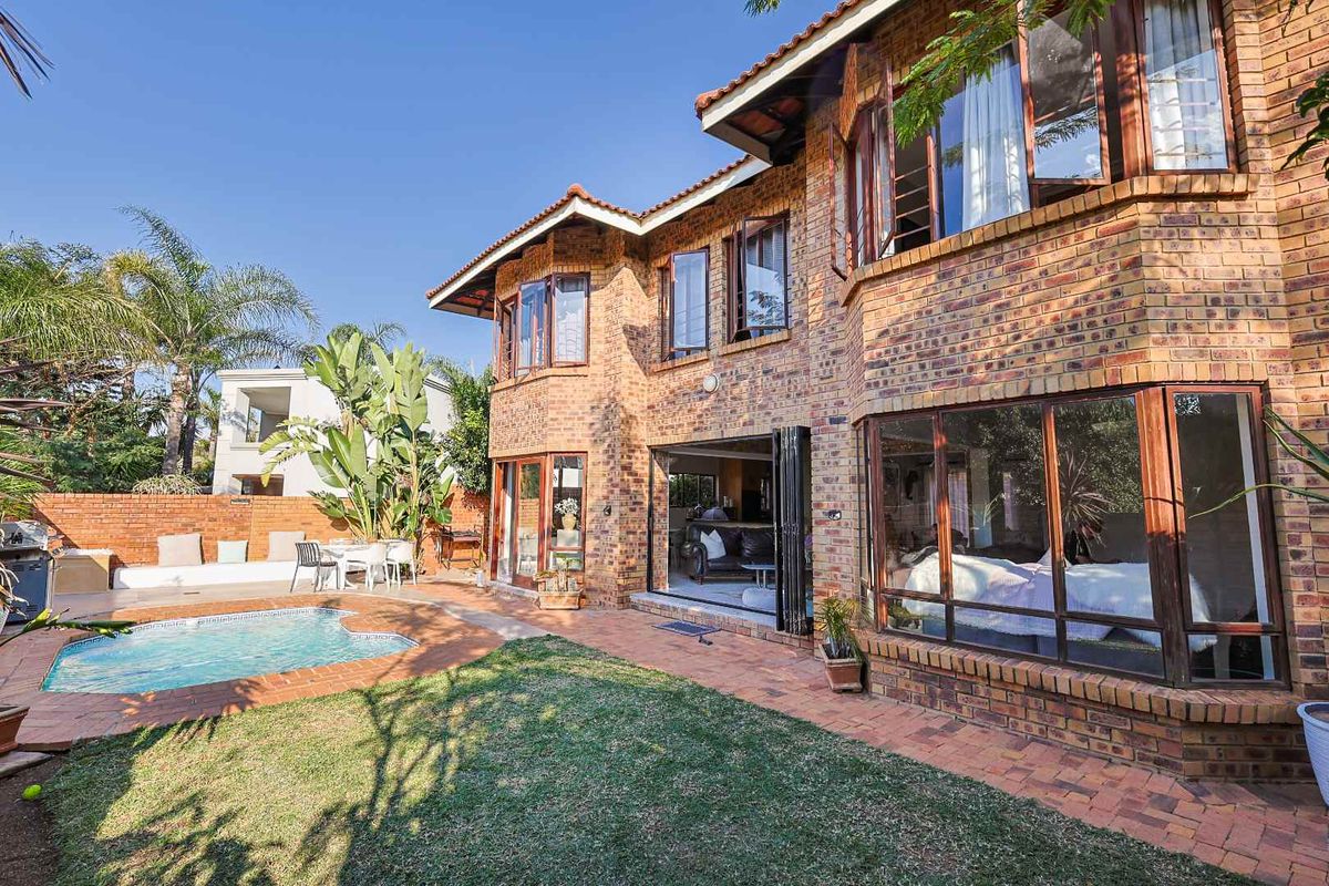 Rear view of a face brick house featuring a swimming pool, paved patio area, and large timber-framed windows with stacking doors leading to the interior.
