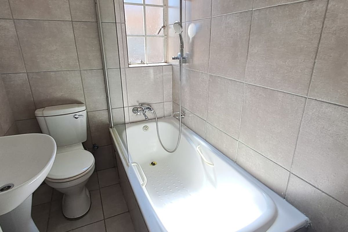 A functional bathroom featuring a pedestal basin, toilet, and a bathtub with a glass screen and handheld shower mixer, finished with neutral wall tiles.