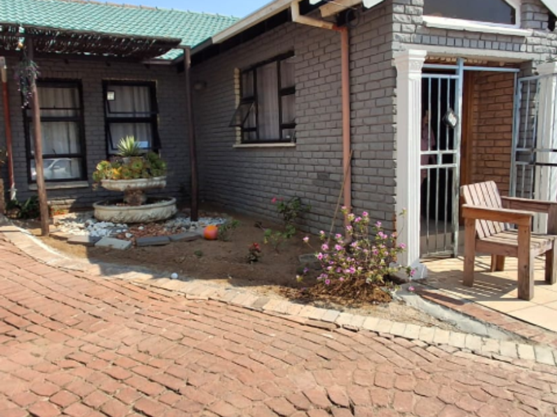 Front exterior view of a brick house featuring a paved driveway, security gate entrance, and a timber pergola over a small garden area.