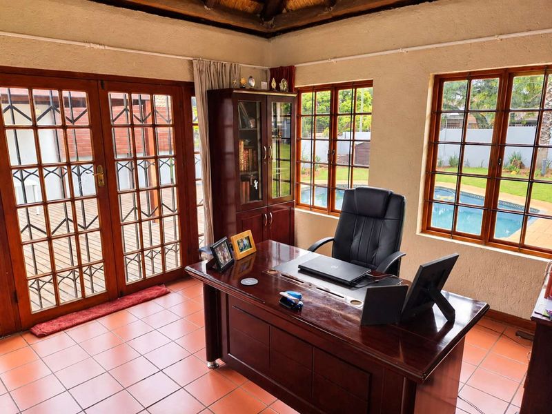A well-lit home office featuring a thatched ceiling, tiled floors, and timber-framed doors and windows with views overlooking the garden and swimming pool.