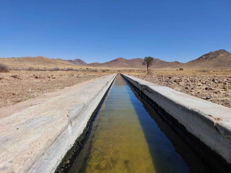 A long concrete water trough situated in an arid farm landscape, featuring mountain views in the background.