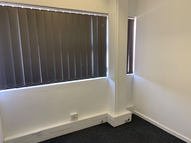 A vacant office space featuring dark carpet flooring, vertical window blinds, and white walls with visible wear on the skirting boards.