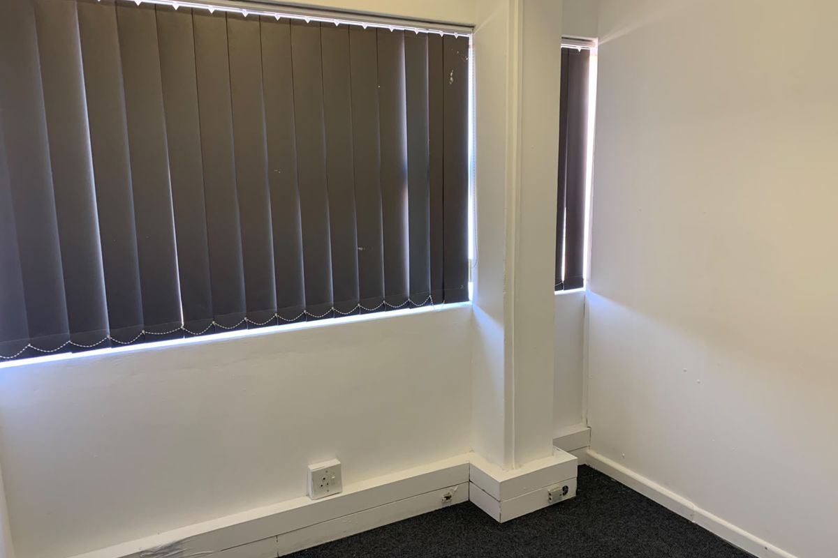 A vacant office space featuring dark carpet flooring, vertical window blinds, and white walls with visible wear on the skirting boards.