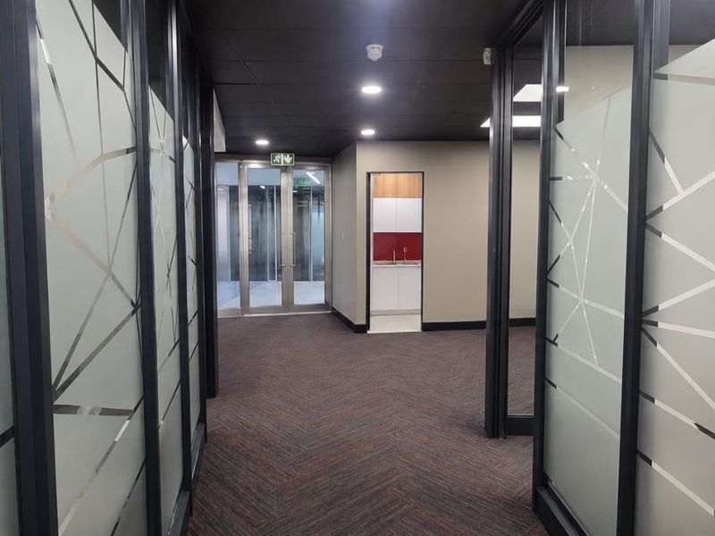 A modern office hallway featuring glass partition walls with geometric frosted detailing, recessed ceiling lighting, and a view toward a small kitchenette area.