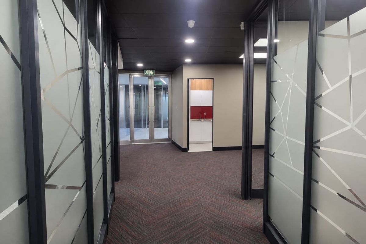 A modern office hallway featuring glass partition walls with geometric frosted detailing, recessed ceiling lighting, and a view toward a small kitchenette area.