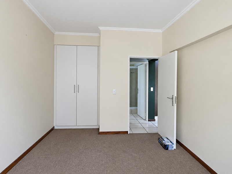 A clean, empty bedroom featuring neutral walls, carpet flooring, and a built-in wardrobe, with an adjacent doorway leading to a tiled bathroom area.