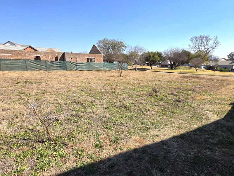 A view of a vacant residential plot featuring dry grass and perimeter fencing, situated in a developed neighborhood.
