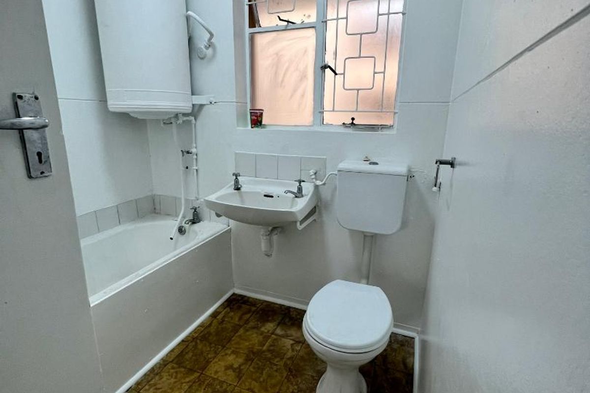 A compact, dated bathroom featuring a bathtub, pedestal basin, toilet, and a wall-mounted electric water heater, requiring modernization of finishes and fixtures.