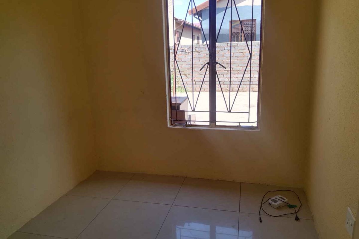 A simple, empty bedroom featuring tiled flooring and a window fitted with security burglar bars.