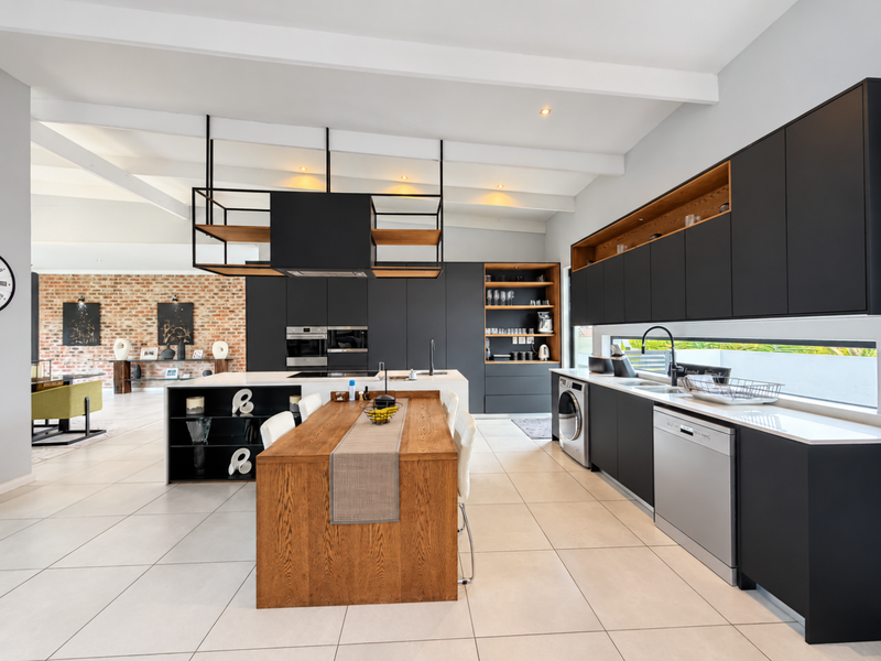 Modern open-plan kitchen featuring sleek matte black cabinetry, a central wooden island, integrated appliances, and striking exposed ceiling beams with an adjacent brick feature wall.