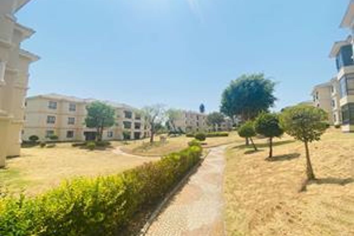 A view of the apartment complex grounds featuring paved walkways, landscaped hedges, and established trees between the residential buildings.