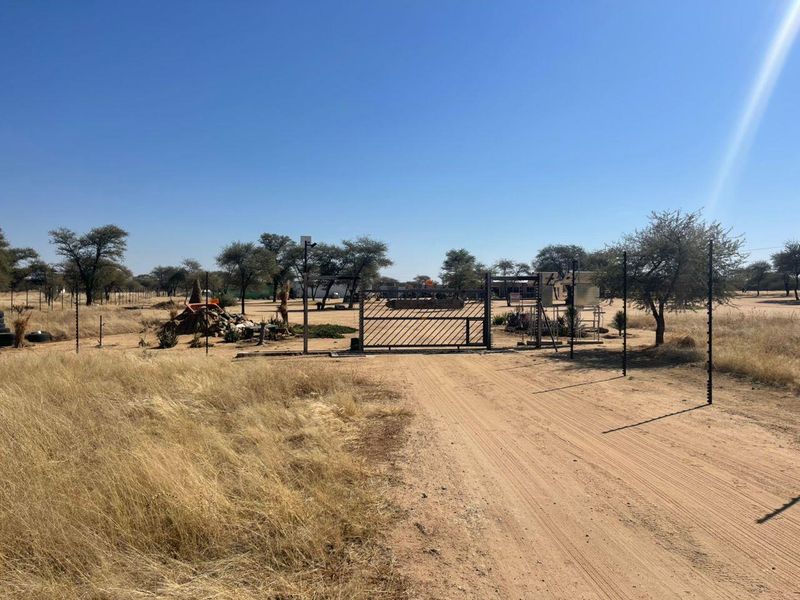A view of the farm entrance featuring a metal security gate, electric fencing, and a dirt driveway leading into the property.