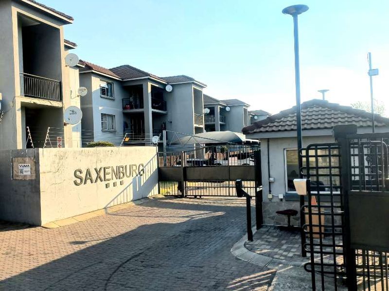 The entrance to Saxenburg apartment complex featuring a paved driveway, security gate, guard house, and multi-story residential buildings with balconies.