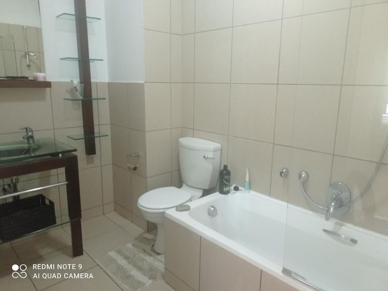 A functional bathroom featuring a glass vessel sink, wall-mounted shelving, a standard toilet, and a bathtub with a glass shower screen and tiled walls.