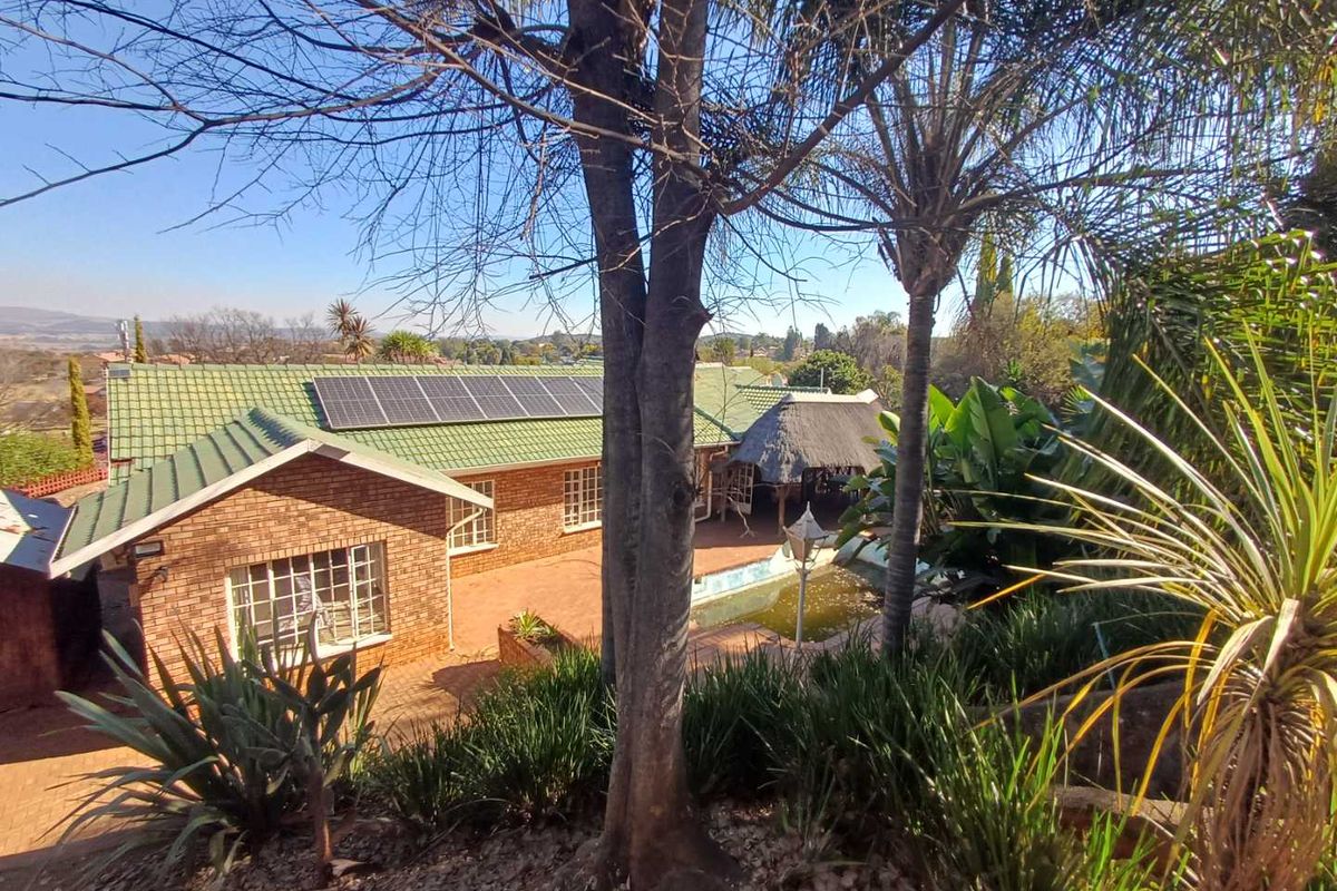 Elevated view of the rear exterior featuring a brick house with a green tiled roof, solar panels, a swimming pool, and a thatched lapa area.
