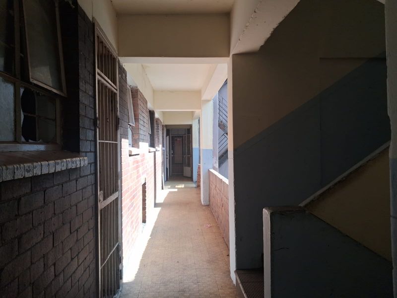 An exterior apartment hallway featuring brick walls, tiled flooring, and a security gate, showing signs of wear and maintenance requirements.