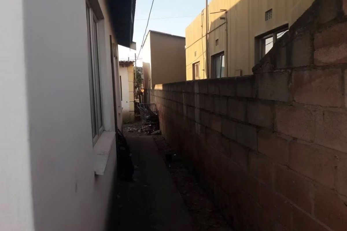A narrow side passage between the property and a concrete block boundary wall, showing signs of wear and debris.