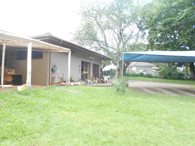 Rear view of the property featuring a covered patio area, paved driveway, and a shade cloth carport structure set within a garden.