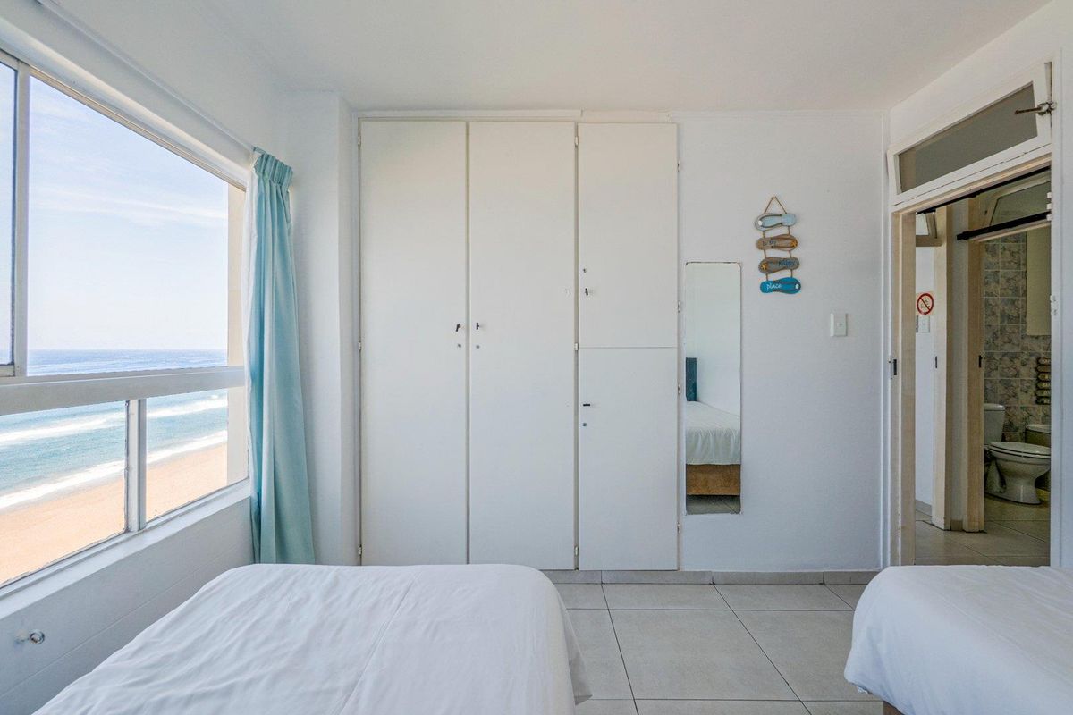 Bedroom featuring built-in cupboards, tiled flooring, and large windows offering direct ocean views, with an adjacent en-suite bathroom.