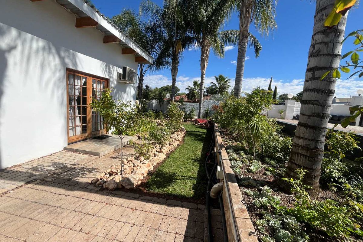 A well-maintained rear garden area featuring paved walkways, established palm trees, and direct access to the guest house via wooden French doors.