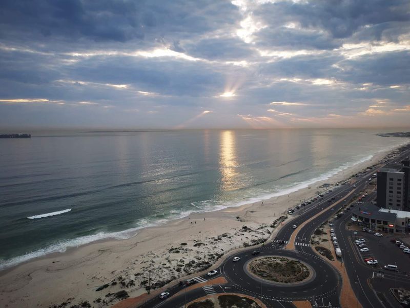 Elevated panoramic view overlooking the coastline, beach, and surrounding road infrastructure under a dramatic cloudy sky.