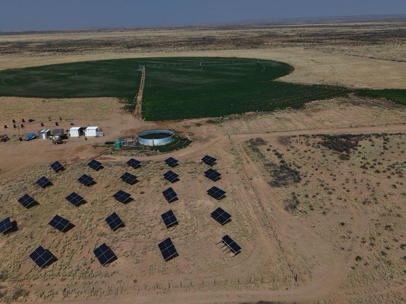 Aerial view of a farm featuring a solar panel array, a circular water reservoir, and a large center pivot irrigation system in the fields.