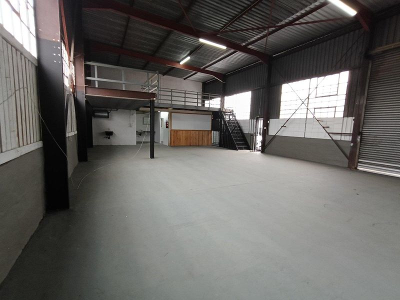 Spacious industrial warehouse unit featuring a mezzanine level, steel staircase, roller shutter door, and concrete flooring, suitable for various commercial or storage applications.