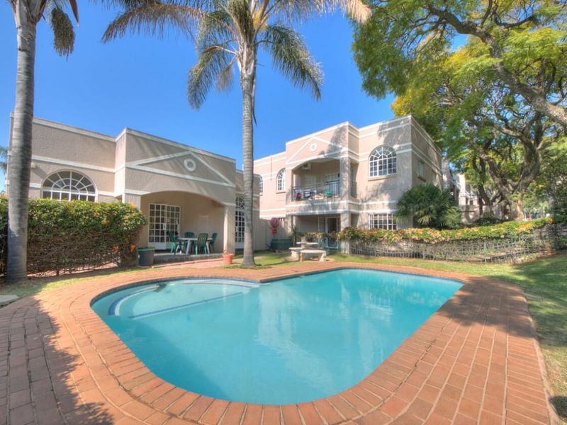A well-maintained apartment complex exterior featuring a central swimming pool with brick coping, surrounded by landscaped gardens and private patio and balcony spaces.