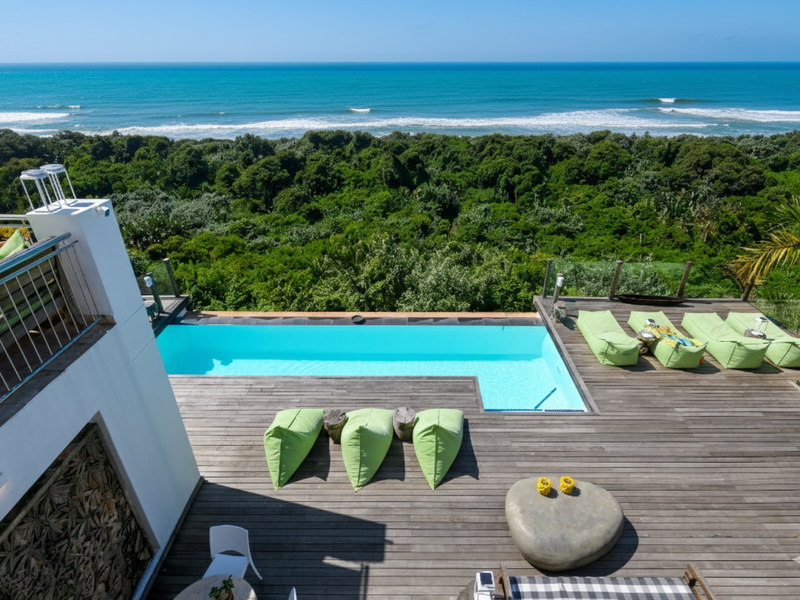 An elevated outdoor pool area featuring timber decking, a modern swimming pool, and a glass balustrade overlooking a lush forest and the ocean.