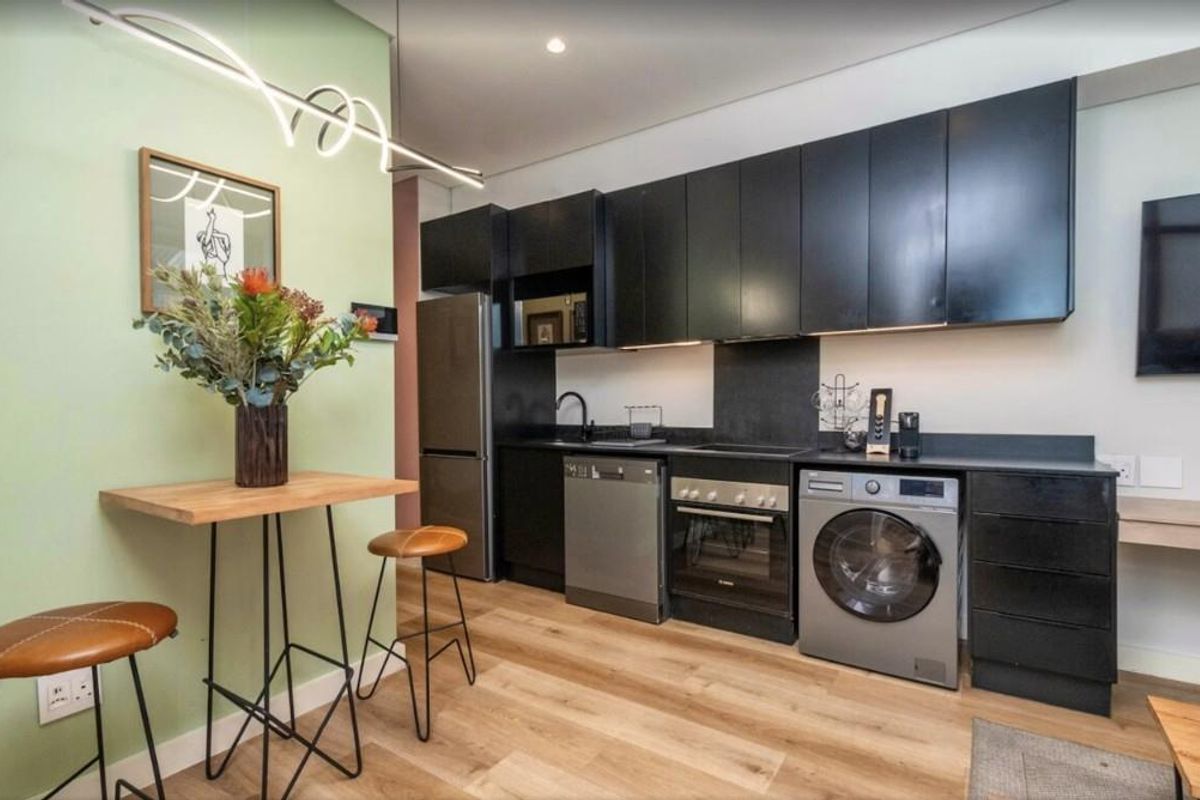 Modern compact kitchen featuring sleek black cabinetry, integrated oven, hob, and built-in laundry appliances, finished with light wood-look flooring.