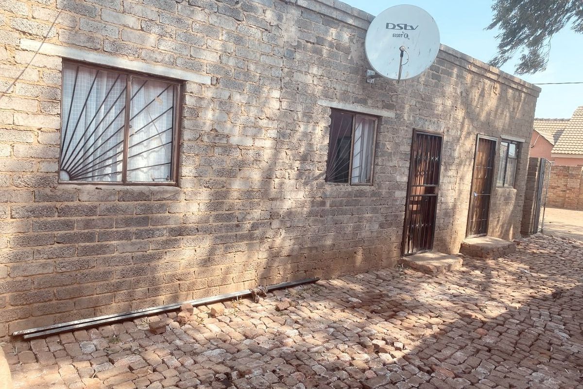Exterior view of a brick-walled residential structure featuring security bars on windows and a satellite dish, set above a cobblestone paved area.