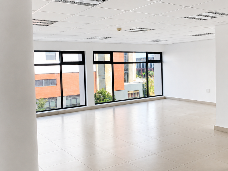 A bright, open-plan office space featuring large windows, neutral ceramic floor tiles, and a suspended ceiling with integrated lighting.