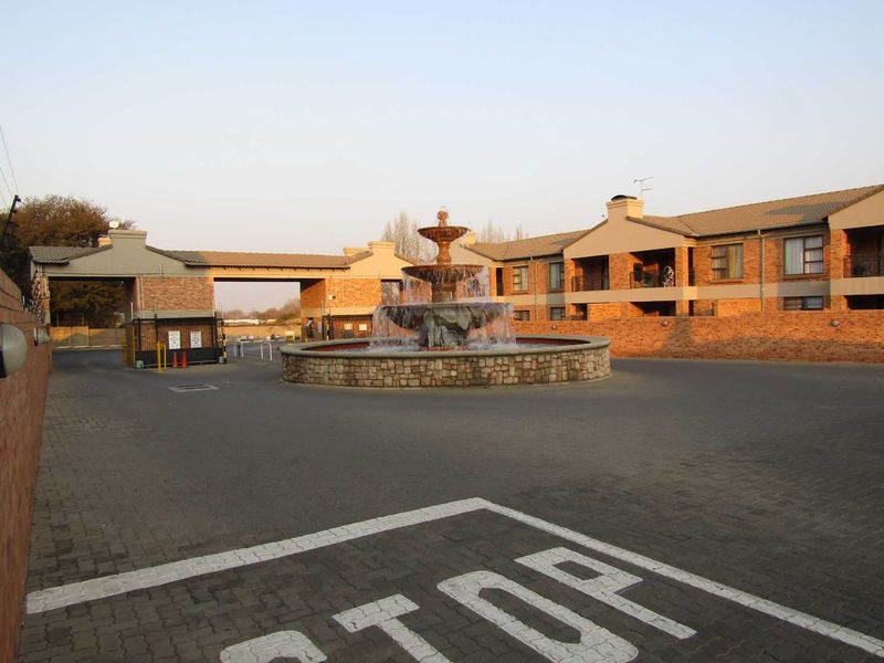 The entrance to the apartment complex features a decorative stone fountain, paved driveway, and secure gated access point.