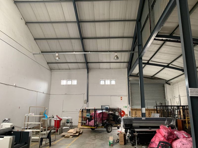 Spacious industrial warehouse unit featuring high ceilings with exposed steel trusses, concrete flooring, and a large roller shutter door for easy access.