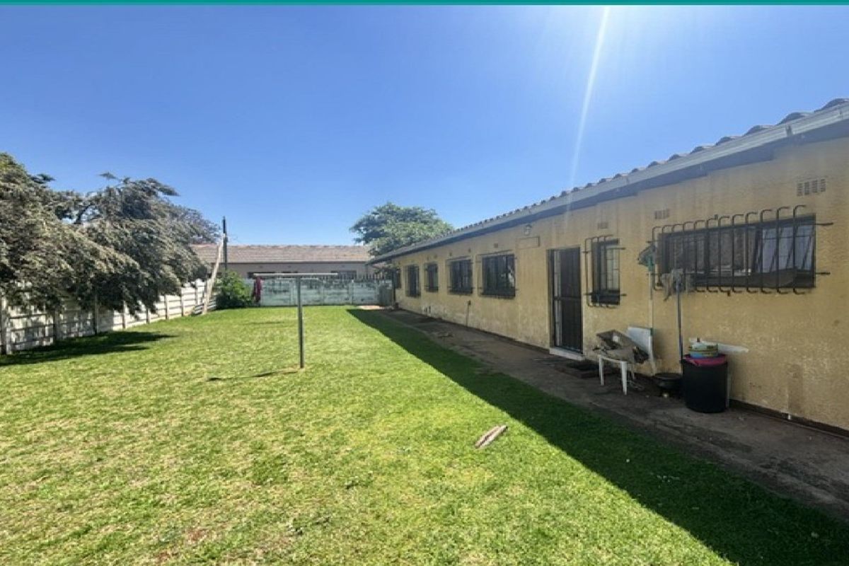 Rear exterior view of the house featuring a large lawn, paved walkway, and security bars on windows, with visible wear on the exterior wall finish.