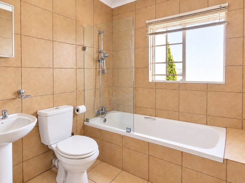 A clean, functional bathroom featuring a pedestal basin, toilet, and a bathtub with a glass shower screen, finished with neutral wall and floor tiles.