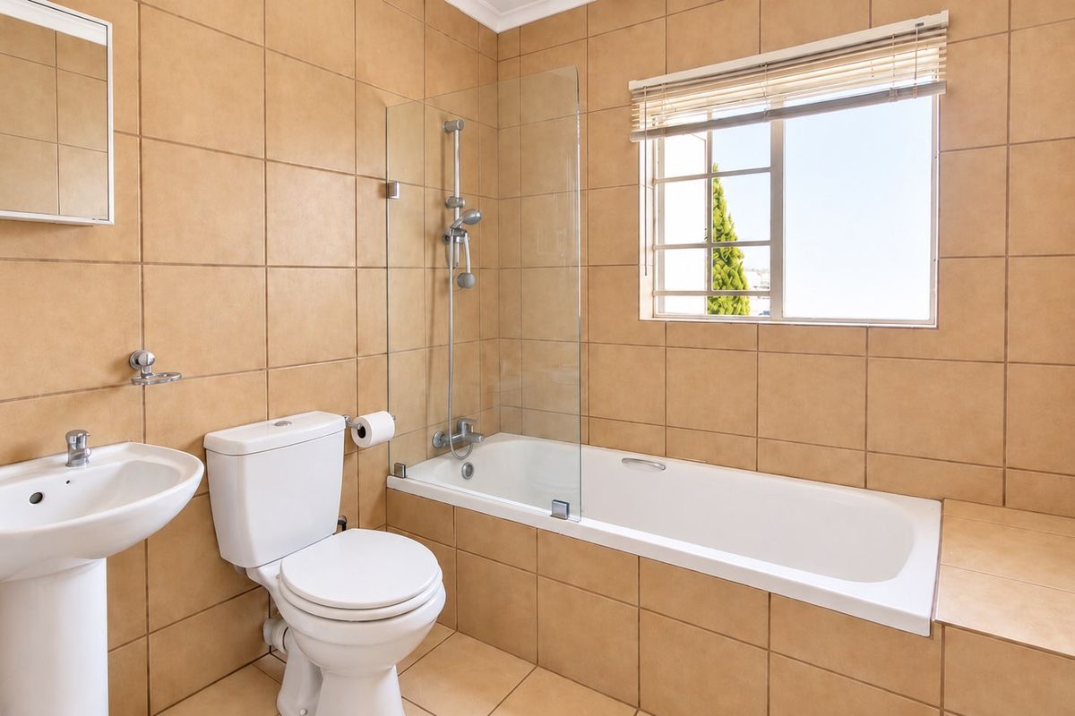 A clean, functional bathroom featuring a pedestal basin, toilet, and a bathtub with a glass shower screen, finished with neutral wall and floor tiles.