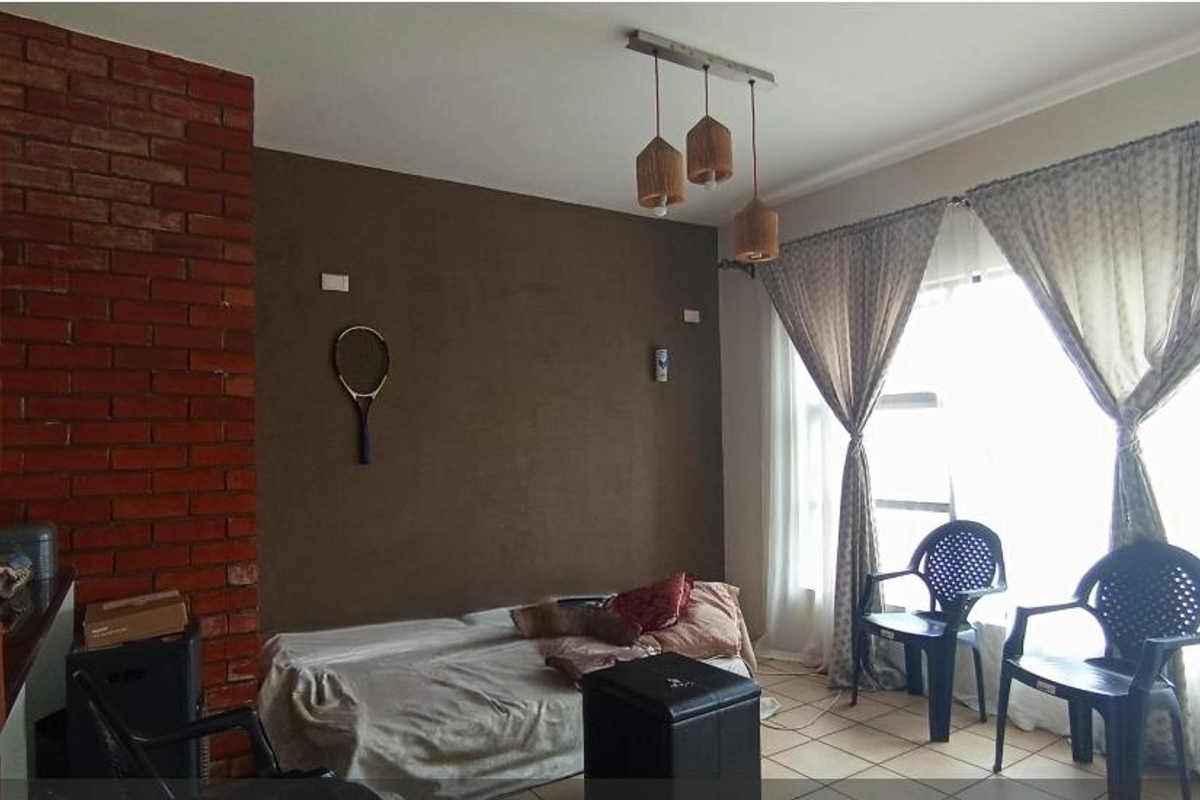 A living room featuring a distinctive red brick accent wall, tiled flooring, and large windows with curtains, providing natural light to the space.