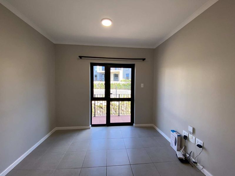 A bright, empty living room featuring neutral tiled flooring and a double glass door providing access to an outdoor area.