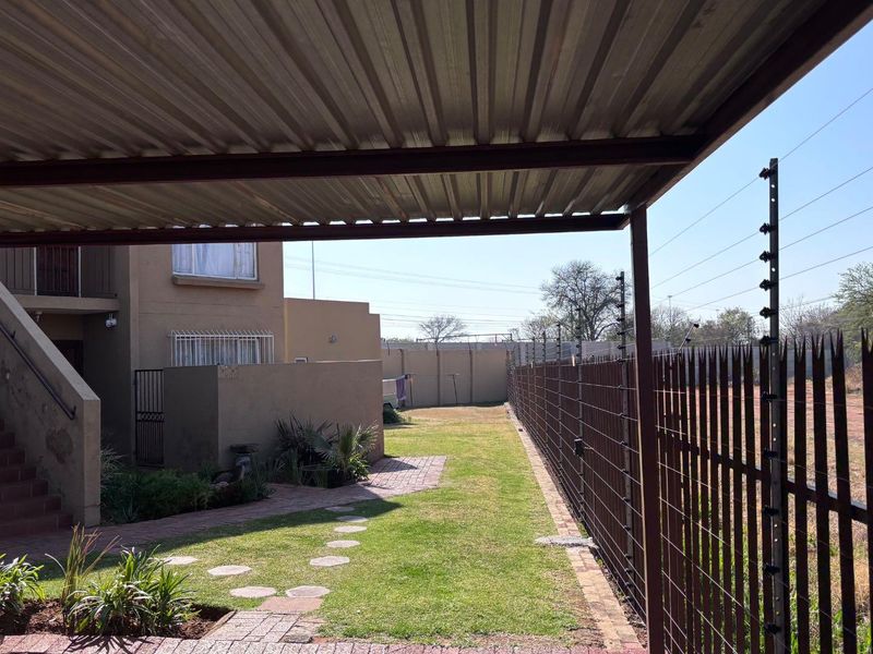 Rear exterior view featuring a covered carport, paved walkway, lawn area, and secure perimeter fencing with electric security wires.