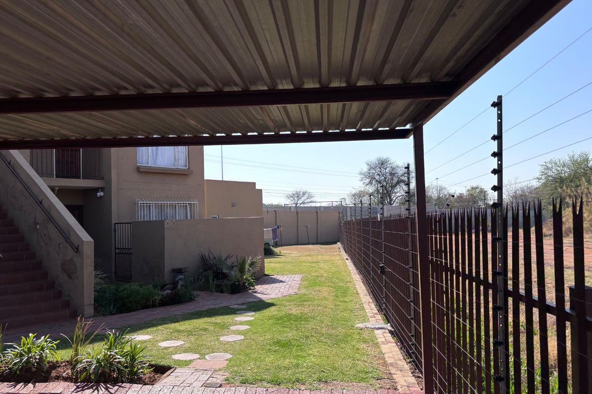 Rear exterior view featuring a covered carport, paved walkway, lawn area, and secure perimeter fencing with electric security wires.