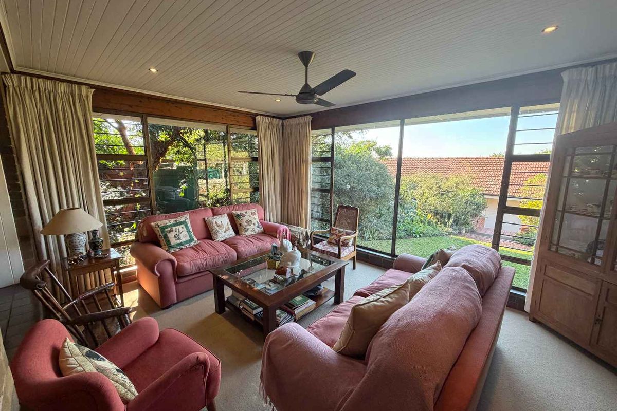 Spacious living room featuring large floor-to-ceiling windows, timber-paneled ceiling, and a ceiling fan, offering abundant natural light and garden views.