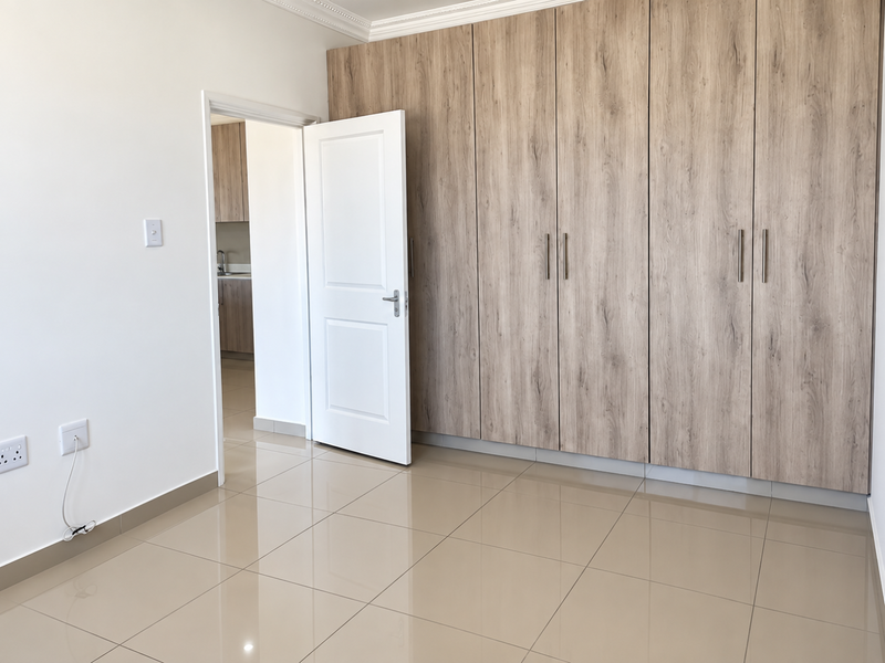 A bright, empty bedroom featuring modern wood-finish built-in cupboards, neutral tiled flooring, and clean white walls with coving detail.