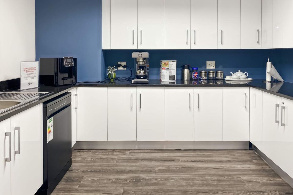 A modern office kitchenette featuring white cabinetry, dark countertops, a stainless steel sink, and integrated dishwasher.